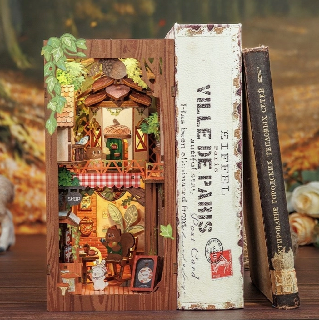 The completed Squirrel Collector Book Nook lit up and placed on a shelf with books. 