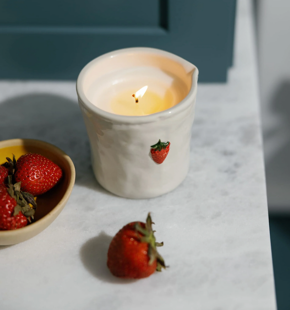 Ceramic candle holder with a lit strawberry  scented candle on a marble surface next to strawberries.