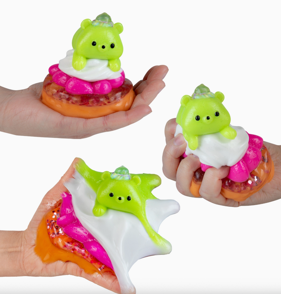 A green bear resting atop a sweet treat held in a hand and being squeezed to show the stickiness and stretchiness of the Taba-Licious Sweetsie. 