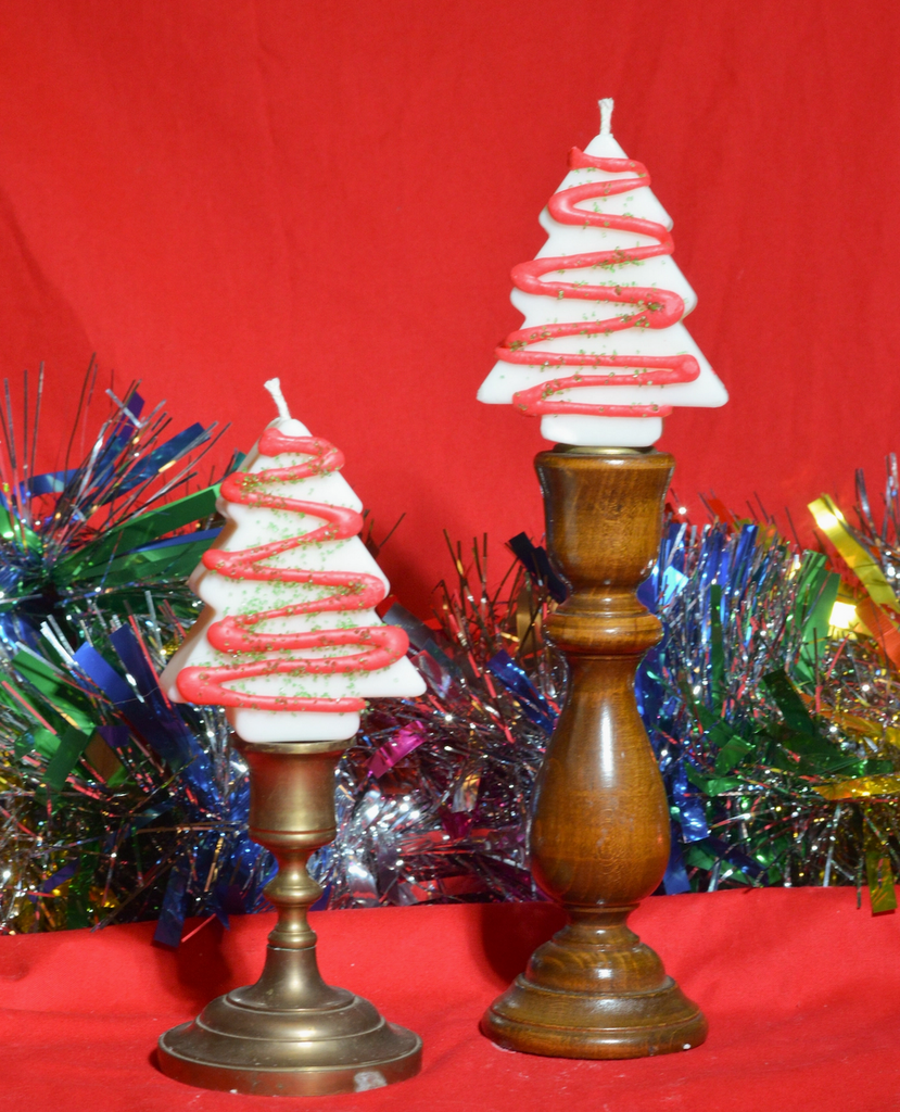 Two Christmas Tree Cake candles with white icing decorated with red swirls and green glitter on wooden stands with a red background and tinsel.
