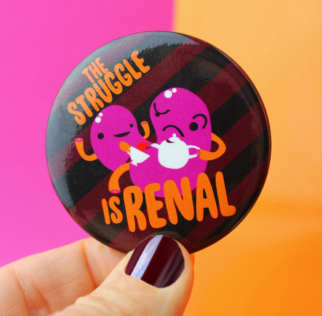 "The Struggle Is Renal" round magnet being held between a thumb and forefinger.
