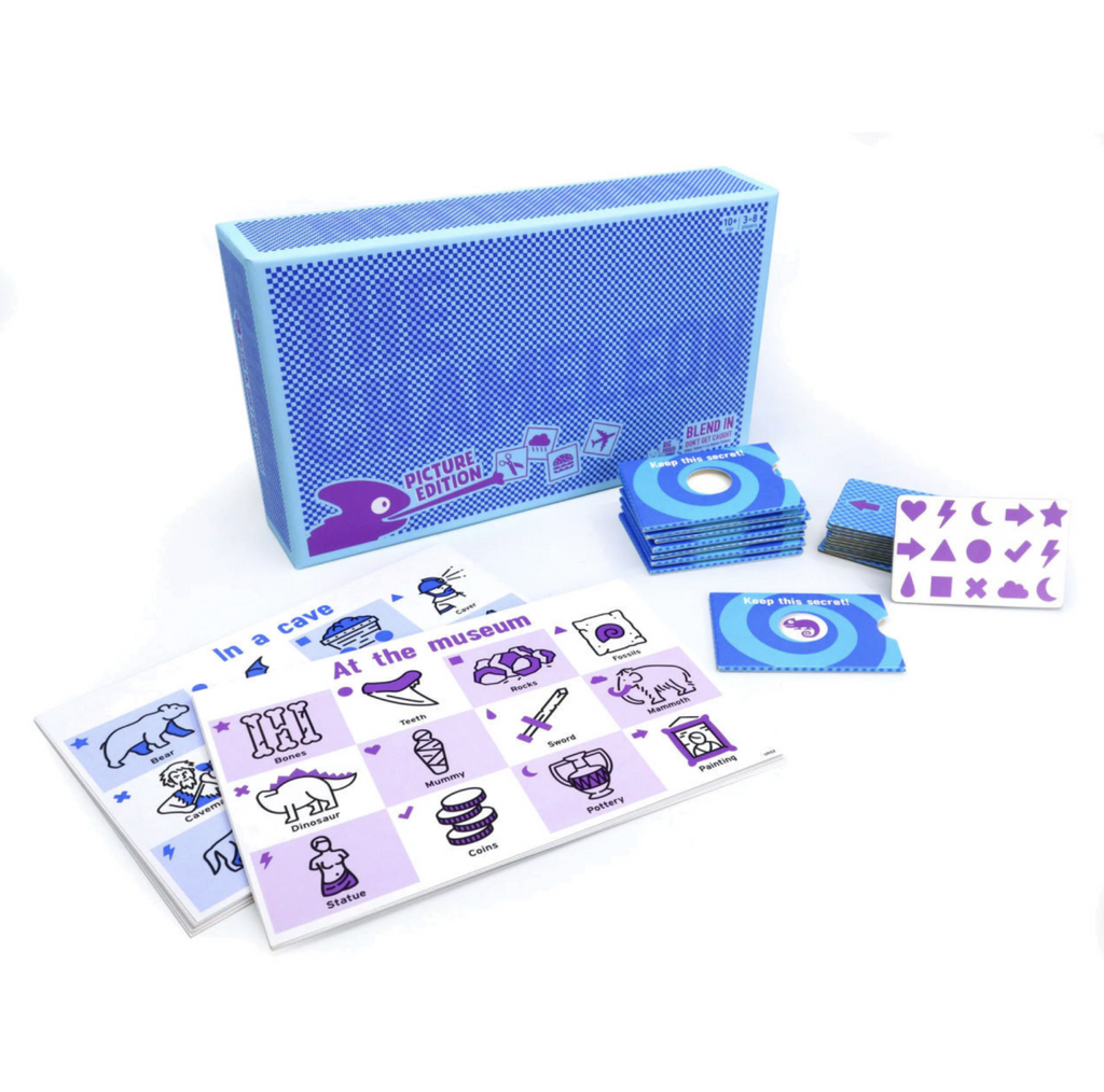 Box and play components of The Chameleon:Picture Edition game. Box is light blue with darker blue dots and a purple chameleon shooting its tongue onto a game card. On table in front of box are game boards, cards, and secret envelopes.