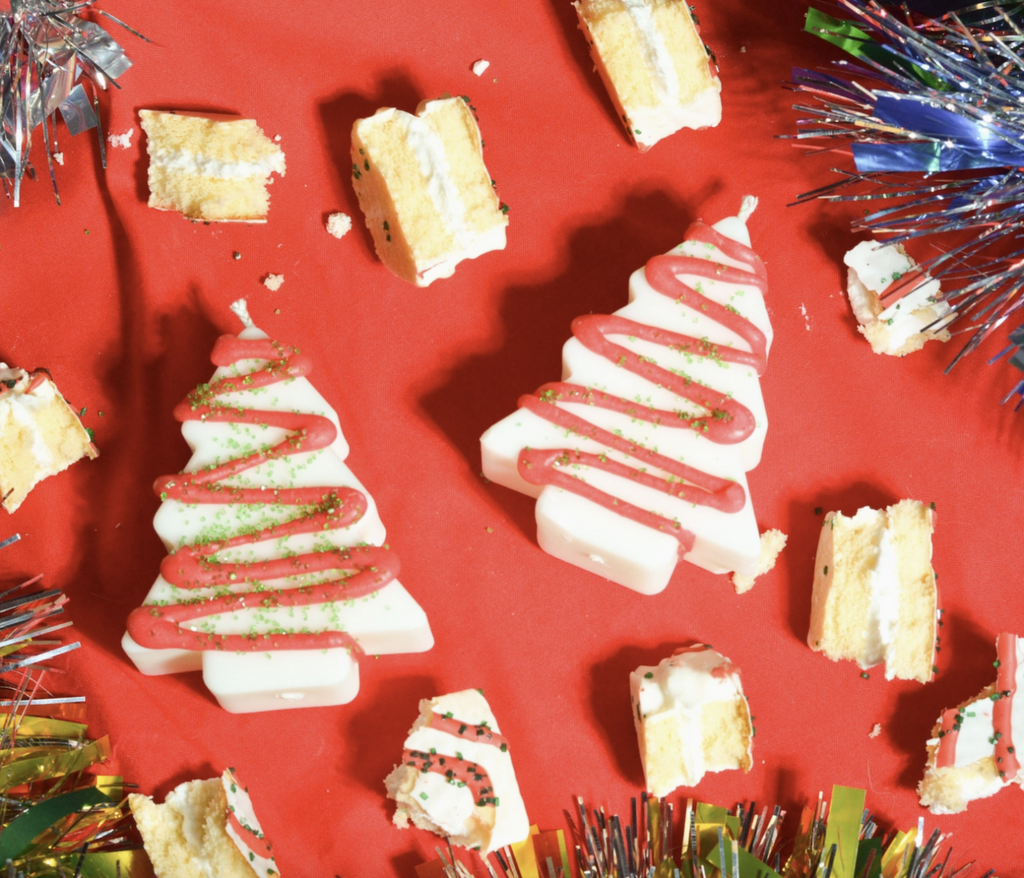Christmas Tree Cake shaped and scented candles laying among actual pieces of Christmas Tree Cake on a red tablecloth. 