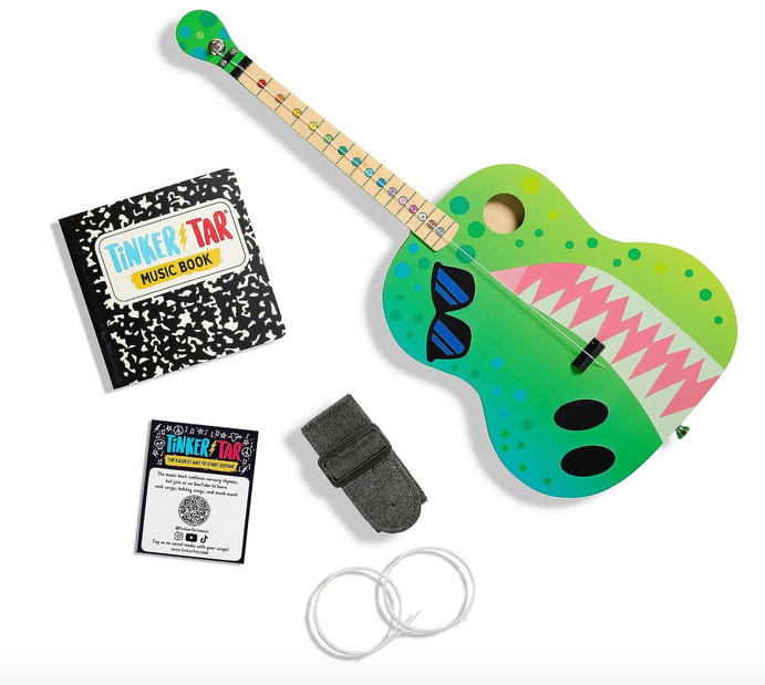 Everything included with the TinkerTar Dino guitar. The guitar painted like the face of a dino, instruction book, shoulder strap, and extra strings. 
