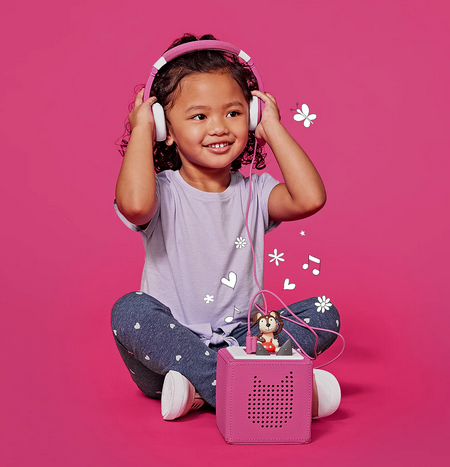 A young child wearing the pink headphones plugged into the pink Toniebox. 