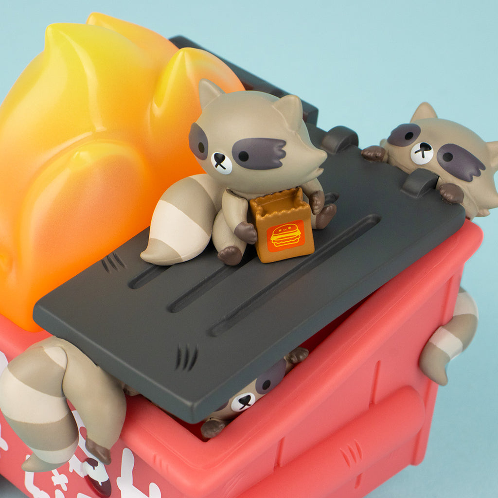 Close up view of Trash Panda Dumpster Fire vinyl figure from the top. We see a panda with a fast food bag sitting on top of the dumpster, another one crawling up the back of the dumpster and a tail sticking out from under the lid. 
