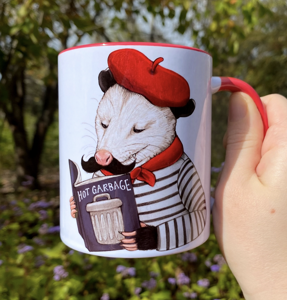 White ceramic mug with red handle and interior with a wonderful illustration of an opossum wearing a black and white striped shirt, red beret and neck kerchief reading a book entitled "Hot Garbage"