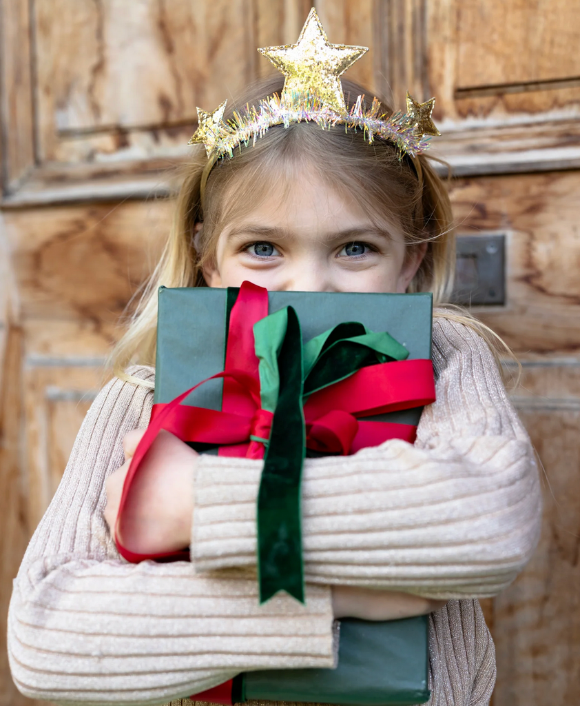 A young child wearing the Twinkle and Tinsel Star Headband with three sparkly gold stars atop a tinsel wrapped headband. She is hugging a festively wrapped present and partially covering her face. 