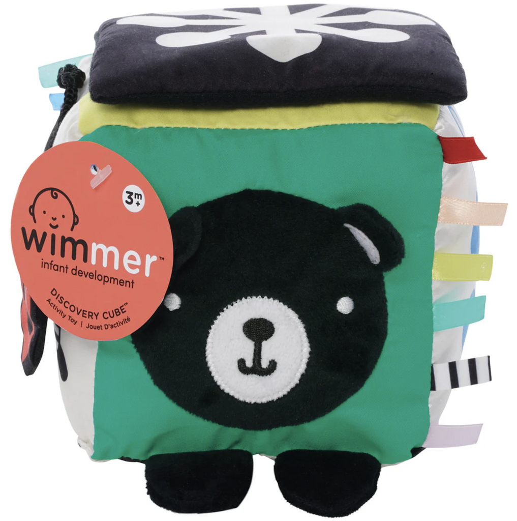 Wimmer Discovery Cube a soft cube with  a baby-safe mirror under a soft fabric flap, textured fabrics, and colored ribbons. A plush bear face with floppy ears and textured nose on a green background is pictured here. 