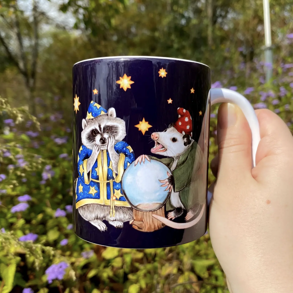 Dark blue mug with a colorful illustration of a fortune telling raccoon with his apprentice opossum around a crystal ball. 