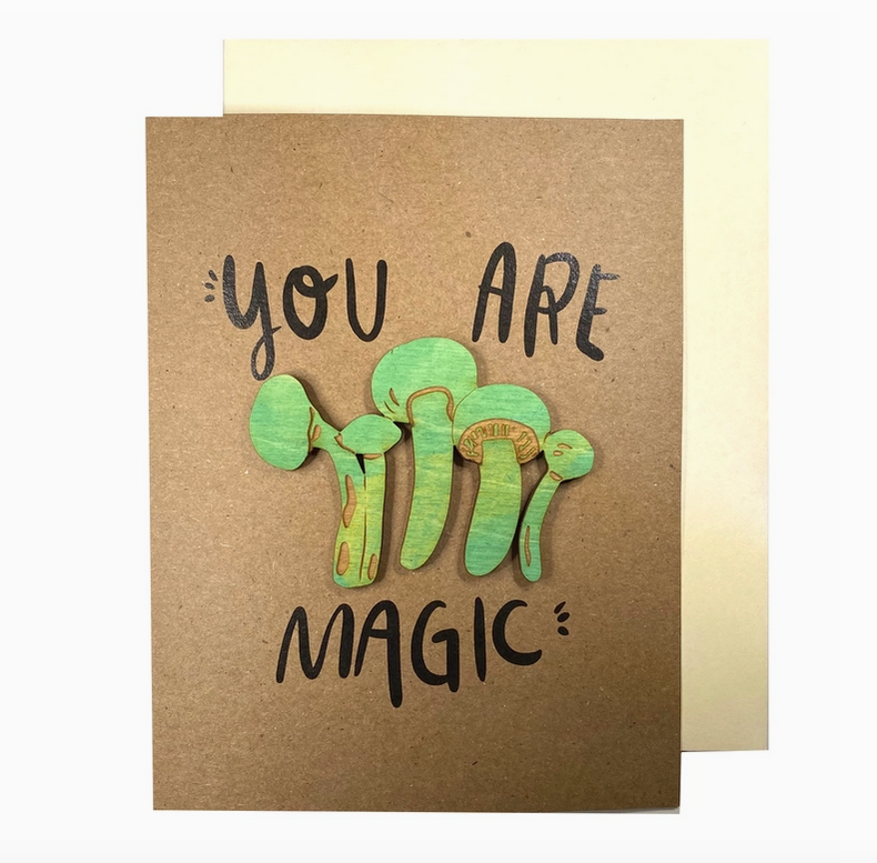 Kraft paper colored greeting card that reads "You Are Magic" in black lettering with greenish yellow, long, skinny mushroom magnets