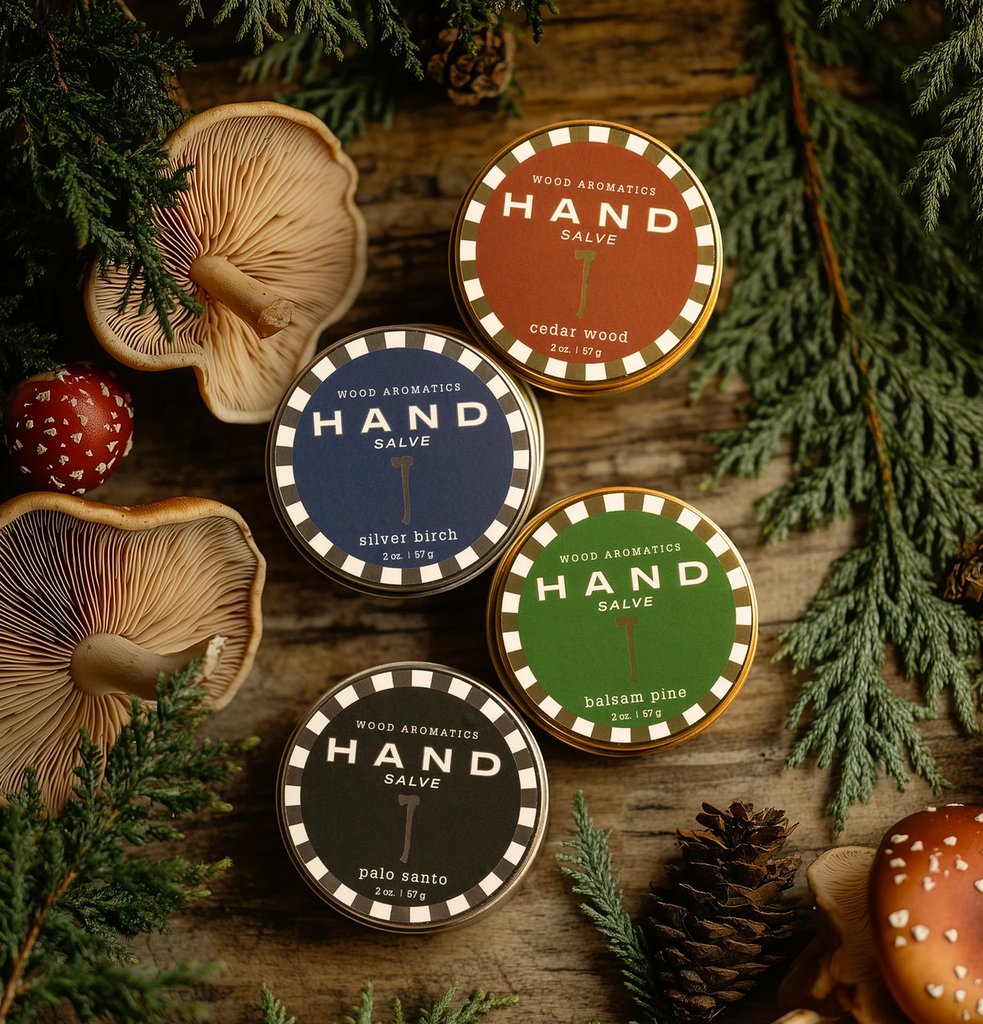 Assorted styles of hand salve tins on a wood table surround by pine branches, pine cones, and mushrooms.