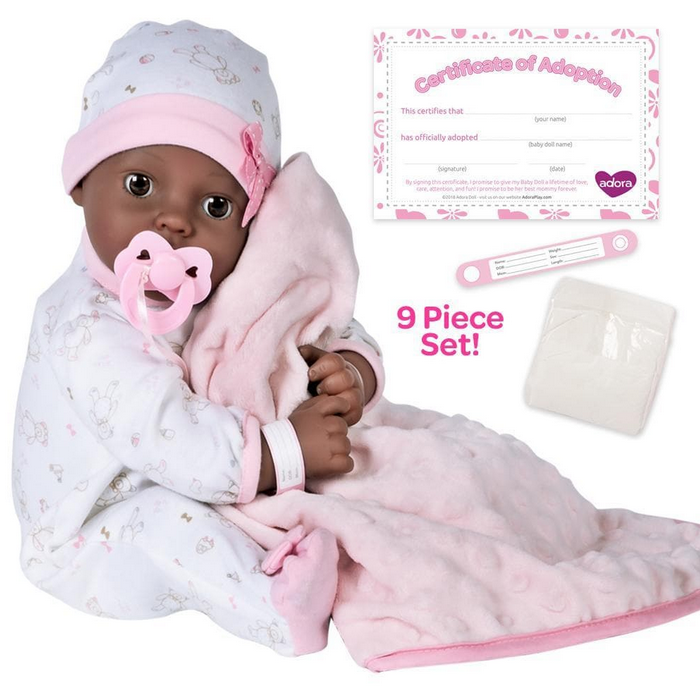 The Joy baby doll set pictured with the 16" weighted baby doll, a Certificate of Adoption, a pacifier, a hospital bracelet, a disposable diaper, and pink blanket. She is sitting up with the pacifier in her mouth.