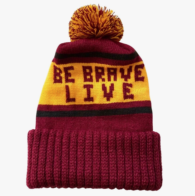 Reddish orange beanie with a yellow band that reads "Be Brave Live" and a yellow and red pom pom on top. 