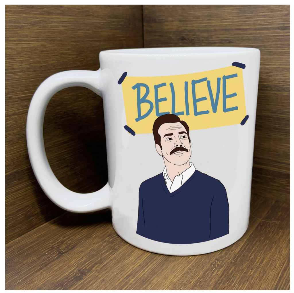 White ceramic mug with a drawing of Ted Lasso under a "Believe" sign.