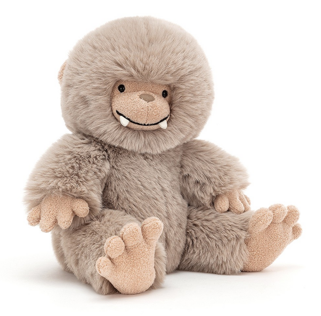 Bo Bigfoot plush sitting upright with his light colored fur and his fangs peeking out from his smiling face. 