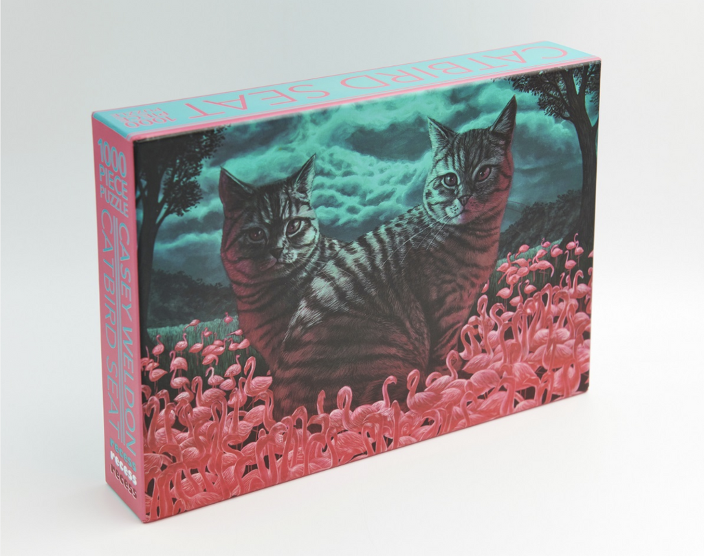 Front of box of Catbird Seat 1000 piece puzzle. Image features two cats sitting in a field of mini pink flamingos.