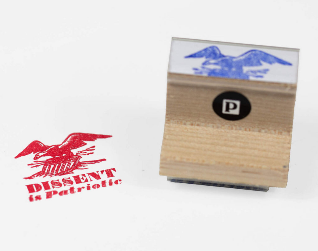 Wooden rubber stamp which reads "Dissent is Patriotic" under an eagle holding a shield that has been pierced with arrows.