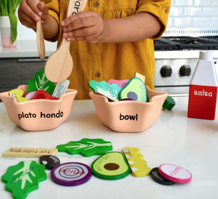 Wooden salad in bowls with dressing and utensils. Each ingredient is labeled in English and Spanish.