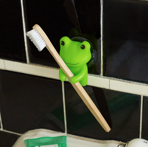 Green silicone frog atatched to a black tile wall and holding a toothbrush. 