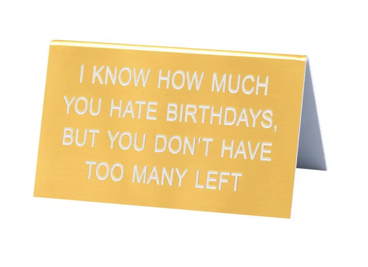 Yellow desk sign reads I know how much you hate birthdays but you don't have many left in white text.