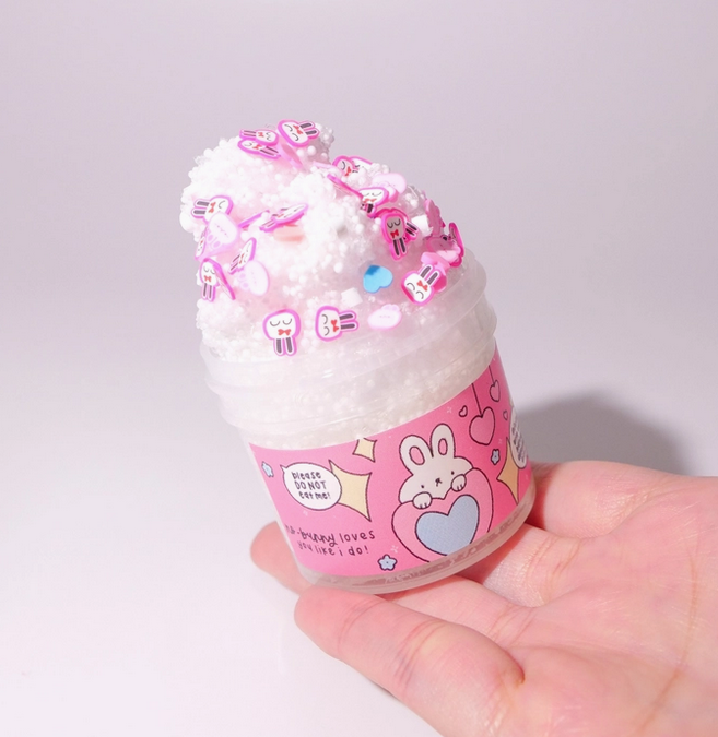 Jar of mini slime with pink label that has illustrated white bunnies and hearts all over it. The slime is fluffy and white with little pink bunny head sprinkles. 