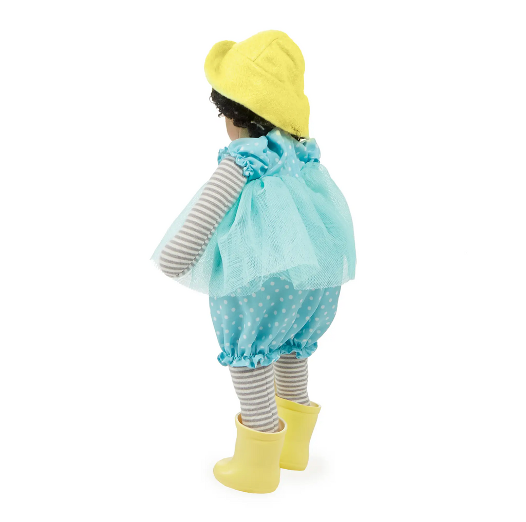 Plush brown skinned doll with curly brown hair. Phoebe is wearing a blue and white polkadot jumpsuit with a blue tulle skirt, a yellow felt hat, and yellow rain boots. backside.