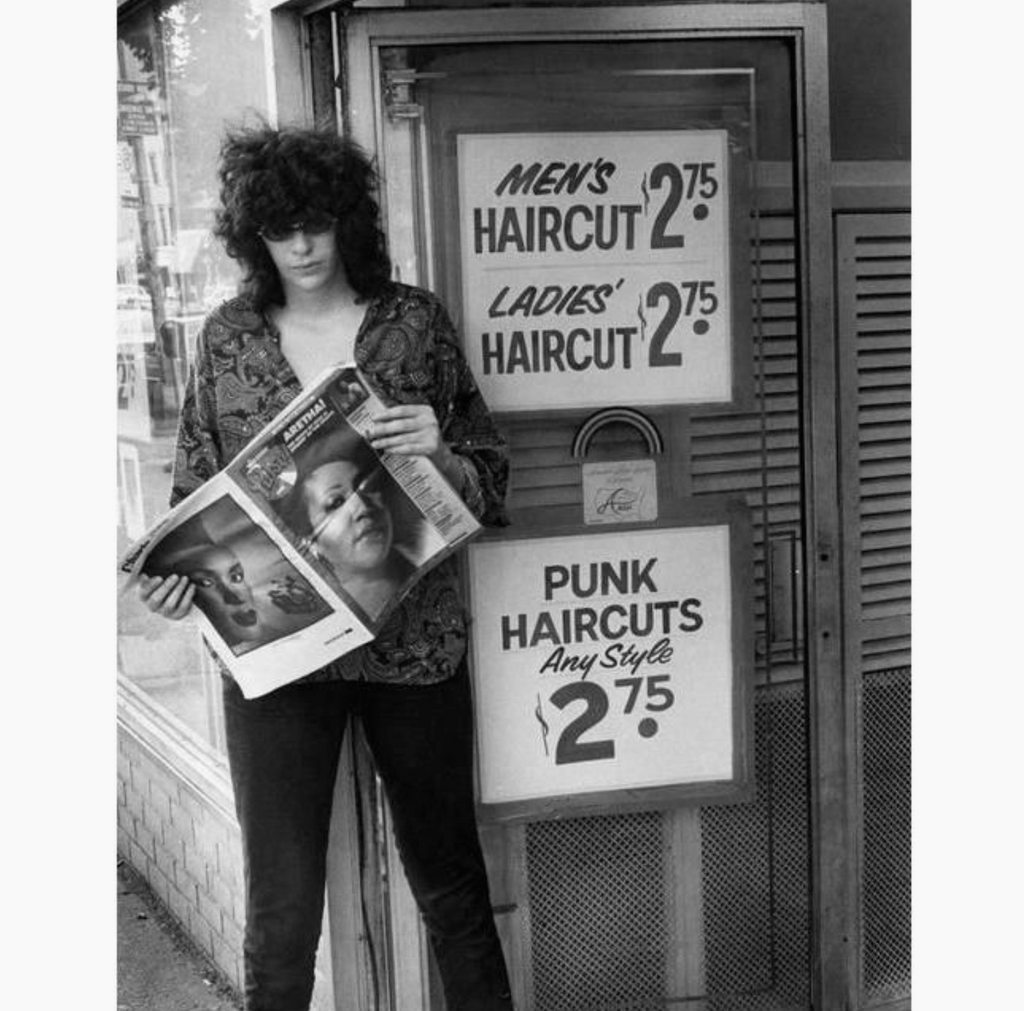 Joey Ramone of The Ramones holding a newspaper next to a sign that reads Punk haircuts Any style $2.75.