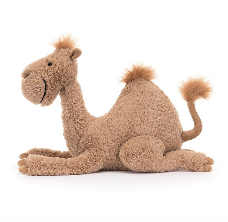 Richie Dromedary plush laying down and viewed from the side. His fur is sandy brown and fluffy. He has a cute little tuft at the top of his head, hump and the end of his tail. 