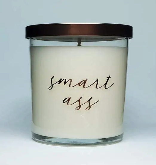White candle in glass that reads "smart ass" in gold cursive.