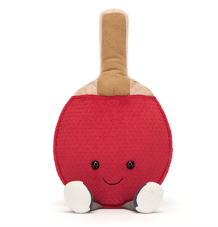 Front of the plush table tennis paddle that has a tan hhandle, red paddle surface, the Jellycat smile and white soled trainers on it's feet. 