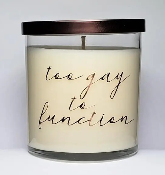 White candle in glass that reads "too gay to function" in gold cursive.