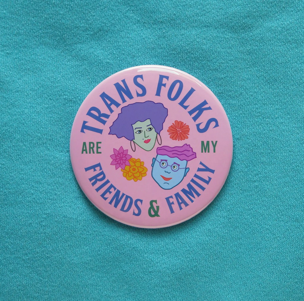 Round 3 inch pinback button that reads Trans Folks are my Friends and Family, with two illustrated faces and flowers on a purple pink background. This picture has the pin alone on a turquoise blue background. 
