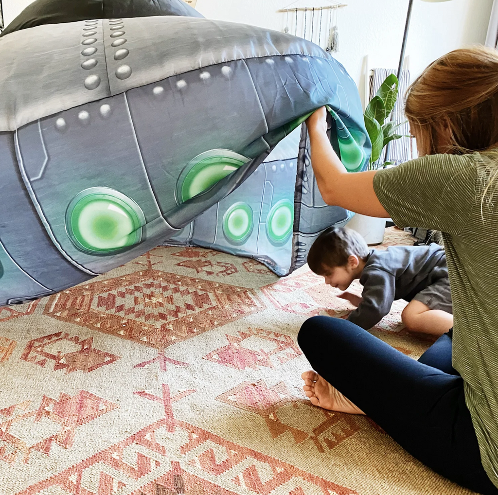 Parent seated on floor pulling up side of inflated UFO Airfort indoor tent for a child to enter.